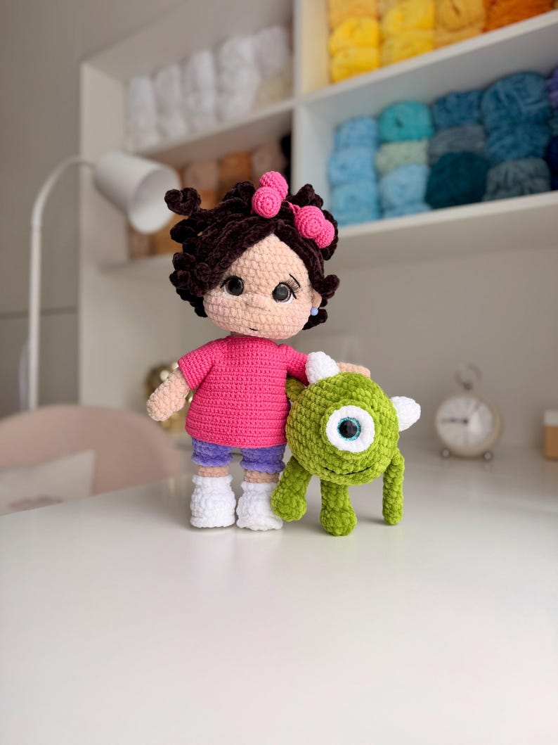 May include: A handmade crochet doll with dark brown curly hair adorned with two pink bows, wearing a bright pink t-shirt, purple shorts, and white shoes. Beside the doll stands a small, round, green one-eyed monster crochet plush toy.