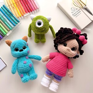 May include: Three handmade crochet dolls including a green one-eyed monster, a blue spotted monster with horns, and a doll with dark curly hair wearing a pink shirt and purple shorts, accompanied by a set of colorful markers and a notebook.