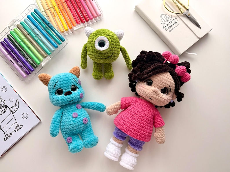 May include: Three handmade crochet dolls including a green one-eyed monster, a blue spotted monster with horns, and a doll with dark curly hair wearing a pink shirt and purple shorts, accompanied by a set of colorful markers and a notebook.