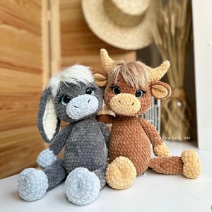 May include: Two crocheted stuffed animals, a gray donkey and a brown cow, are sitting together on a white surface. The donkey has a white face and blue eyes, while the cow has brown fur and black eyes. Both animals have a friendly expression.