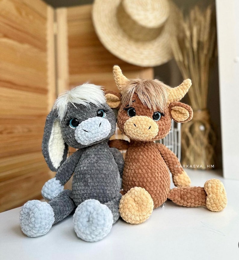 May include: Two crocheted stuffed animals, a gray donkey and a brown cow, are sitting together on a white surface. The donkey has a white face and blue eyes, while the cow has brown fur and black eyes. Both animals have a friendly expression.