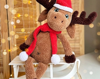 Puede incluir: Un alce de crochet marrón que lleva un gorro de Papá Noel rojo y una bufanda roja, sentado en un trineo de madera blanco. El alce tiene cuernos marrones y pompones blancos en sus pezuñas.