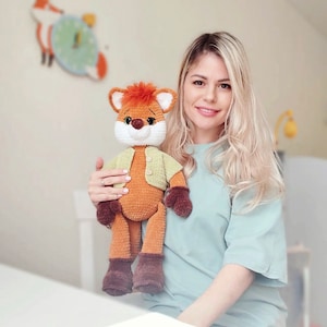 May include: A woman is holding a crocheted fox plush toy. The fox is brown and orange with a green shirt. The woman is wearing a light blue shirt.