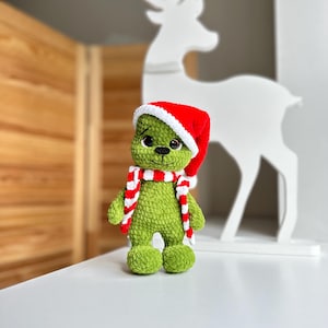 Peut inclure: Une peluche crochet&eacute;e verte &agrave; la surface douce et textur&eacute;e, dot&eacute;e de grands yeux noirs et d'un petit nez noir. La figurine porte un bonnet de P&egrave;re No&euml;l rouge et blanc ainsi qu'une &eacute;charpe ray&eacute;e rouge et blanche assortie autour du cou.