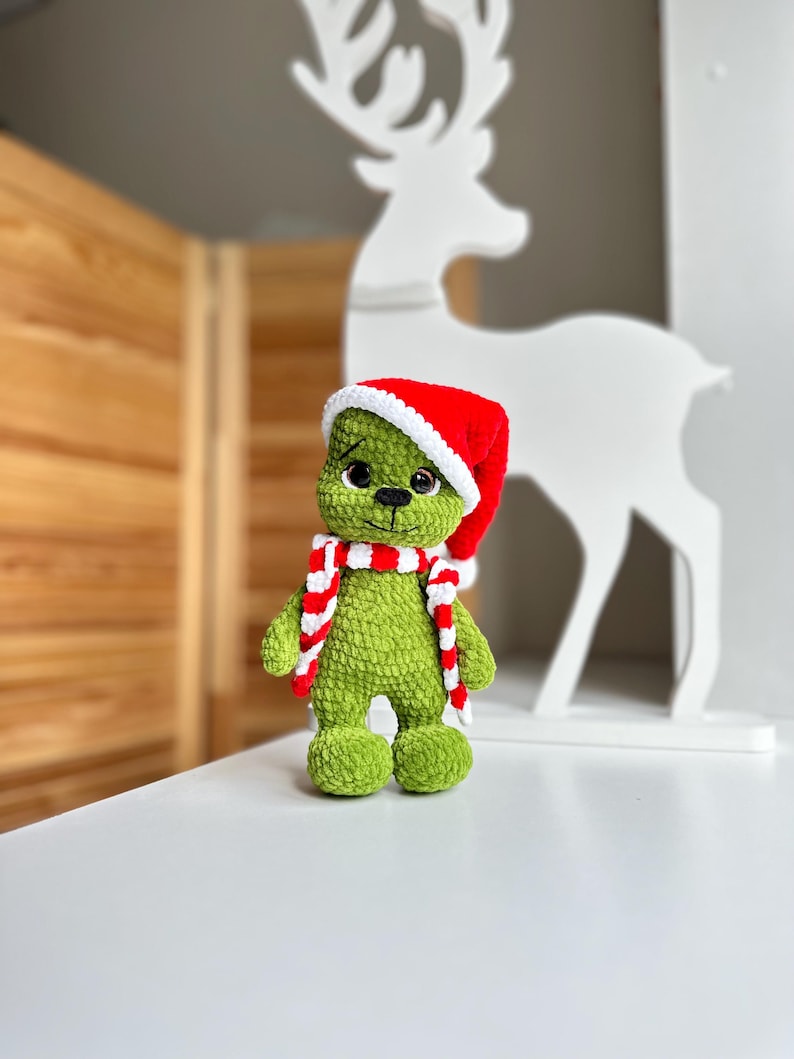 Peut inclure: Une peluche crochet&eacute;e verte &agrave; la surface douce et textur&eacute;e, dot&eacute;e de grands yeux noirs et d'un petit nez noir. La figurine porte un bonnet de P&egrave;re No&euml;l rouge et blanc ainsi qu'une &eacute;charpe ray&eacute;e rouge et blanche assortie autour du cou.