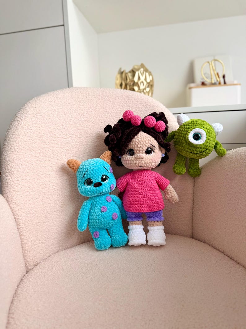 May include: Three handmade crochet dolls: a blue monster with purple spots, a human figure with dark curly hair wearing a pink shirt and purple shorts, and a small green one-eyed monster with white horns, all crafted with yarn.