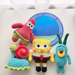 May include: Four crocheted plush toys featuring characters from SpongeBob SquarePants: a pink starfish, a snail with a pink shell, a yellow rectangular sponge, and a small teal creature with one red eye, arranged before a circular blue porthole.