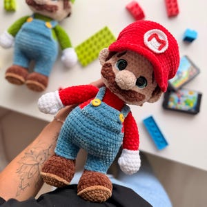 May include: A plush crochet doll of a character wearing a red hat with a white M emblem, a red shirt, blue overalls with yellow buttons, white gloves, and brown shoes. A second similar plush doll with a green hat and L emblem is visible nearby.