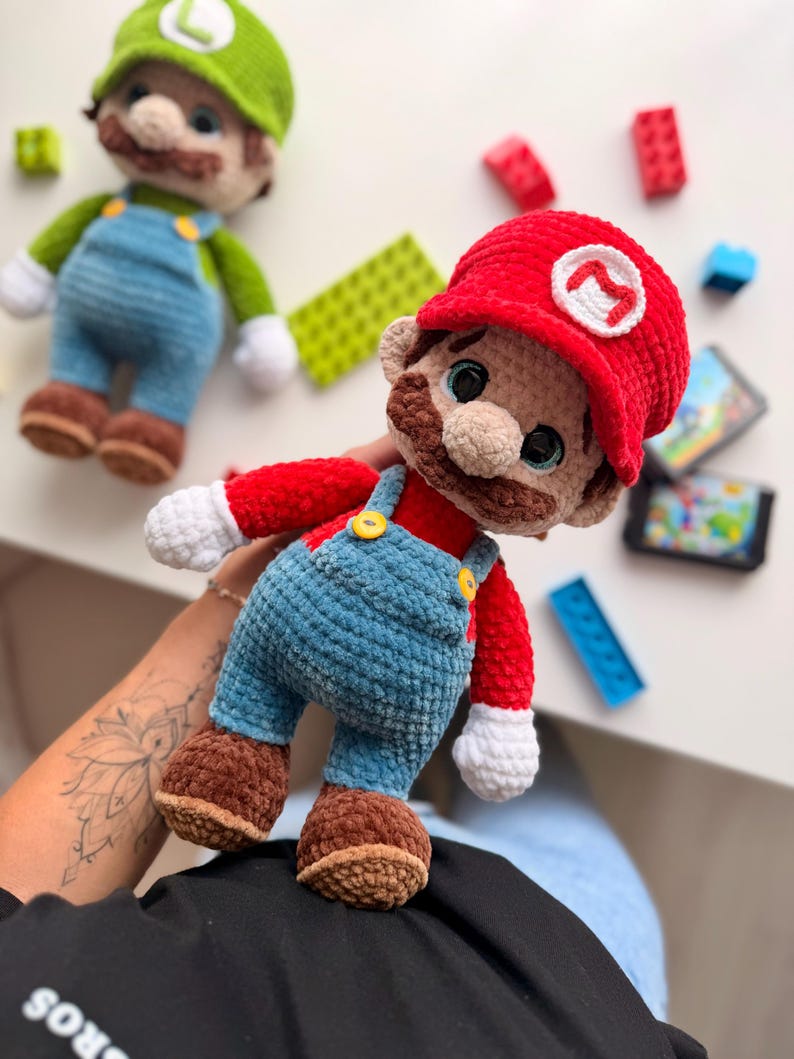 May include: A plush crochet doll of a character wearing a red hat with a white M emblem, a red shirt, blue overalls with yellow buttons, white gloves, and brown shoes. A second similar plush doll with a green hat and L emblem is visible nearby.