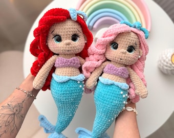 Puede incluir: Dos muñecas de sirena hechas a mano con ganchillo. Una muñeca tiene el pelo rojo, un top morado y una cola azul. La otra muñeca tiene el pelo rosa, un top morado y una cola azul. Ambas muñecas tienen ojos grandes y están decoradas con perlas.