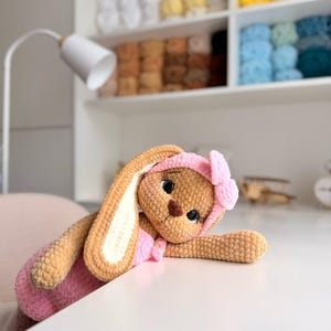 May include: A tan crocheted rabbit plush toy with long ears, wearing a pink headband with a bow and a matching pink outfit. The rabbit has large dark eyes with long eyelashes and a brown stitched nose, crafted from soft, textured yarn material.