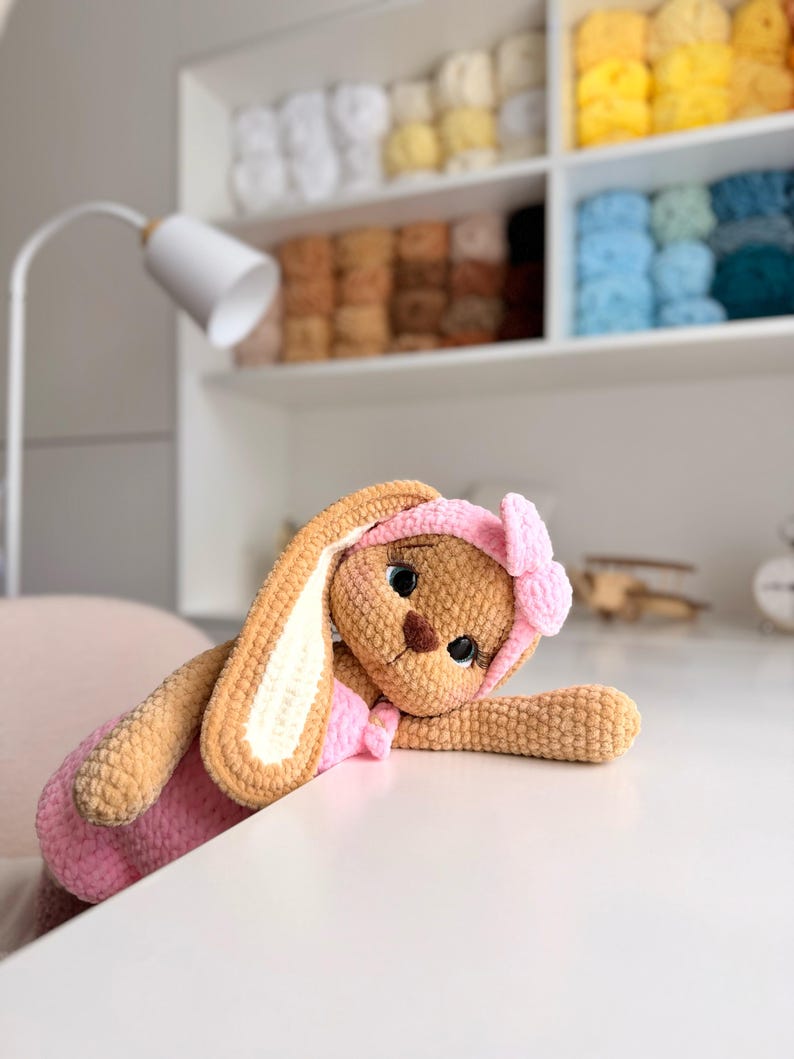 May include: A tan crocheted rabbit plush toy with long ears, wearing a pink headband with a bow and a matching pink outfit. The rabbit has large dark eyes with long eyelashes and a brown stitched nose, crafted from soft, textured yarn material.