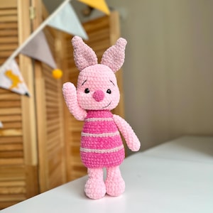 Könnte beinhalten: Ein handgefertigtes Häkel-Plüschtier in Form eines kleinen rosa Schweinchens mit großen Ohren, schwarzen Perlenaugen und einer dunkelrosa Nase. Der Körper ist mit horizontalen Streifen in abwechselnden hellen und dunklen Rosatönen versehen.