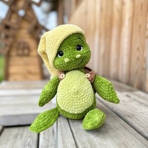 Könnte beinhalten: Ein grünes Häkel-Schildkrötenspielzeug, das eine gelbe Häkel-Schlafkappe trägt. Die Schildkröte hat braune Knopfaugen und eine braune Knopfnase. Das Spielzeug sitzt auf einer Holzoberfläche.