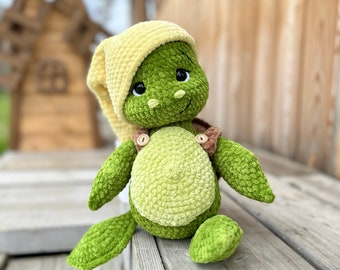 Puede incluir: Un juguete de tortuga tejido a ganchillo con un cuerpo verde texturizado, vientre verde claro y un gorro de dormir amarillo suave. La tortuga tiene ojos negros grandes, una pequeña sonrisa bordada y tirantes marrones con botones de madera.
