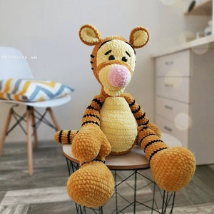 May include: A crocheted stuffed toy tiger with orange and black stripes, a yellow body, and a pink nose. The toy is sitting on a wooden table with a black metal base.