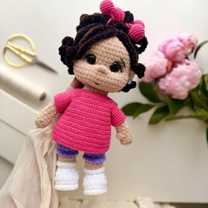May include: A handmade crochet doll featuring dark brown hair styled with pink bows, wearing a bright pink short-sleeved shirt, purple shorts, and white shoes. The doll has large black eyes, a small nose, and a light beige complexion.