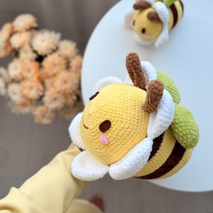 K&ouml;nnte beinhalten: Geh&auml;kelte Bienen-Pl&uuml;schtiere mit gelben K&ouml;rpern, braunen Streifen und wei&szlig;en und gr&uuml;nen Akzenten. Die Bienen haben l&auml;chelnde Gesichter und braune F&uuml;hler. Eine Biene ist im Vordergrund, eine weitere im Hintergrund.