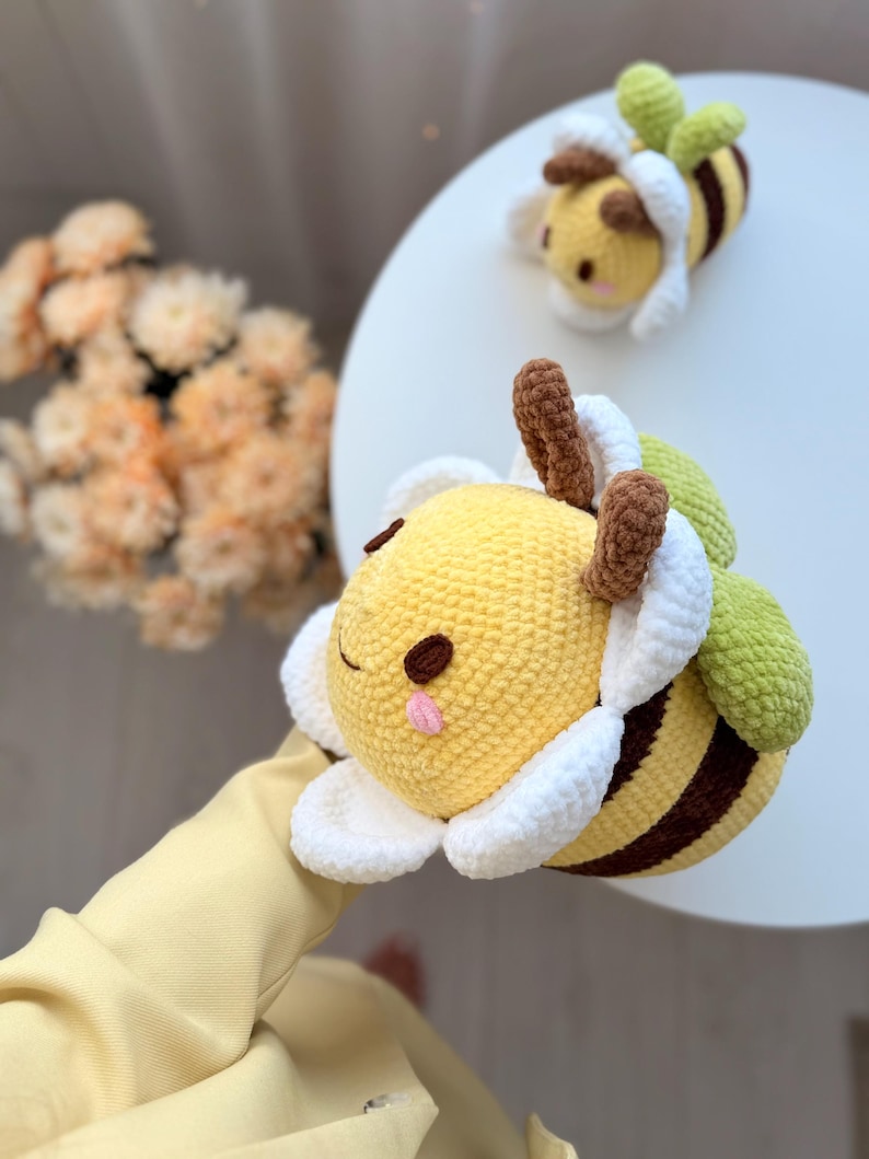 K&ouml;nnte beinhalten: Geh&auml;kelte Bienen-Pl&uuml;schtiere mit gelben K&ouml;rpern, braunen Streifen und wei&szlig;en und gr&uuml;nen Akzenten. Die Bienen haben l&auml;chelnde Gesichter und braune F&uuml;hler. Eine Biene ist im Vordergrund, eine weitere im Hintergrund.