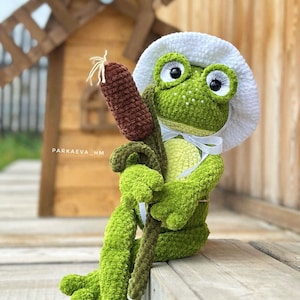 May include: A handmade green crochet frog doll wearing a white hat and holding a brown cattail. The frog has large eyes and is sitting on a wooden surface. The doll is a whimsical, handcrafted item.