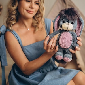 Pu&ograve; includere: Una donna con un vestito blu tiene in mano un peluche di stoffa grigio e rosa lavorato a uncinetto con orecchie lunghe. L'animale ha un viso cucito con occhi neri e un naso nero.