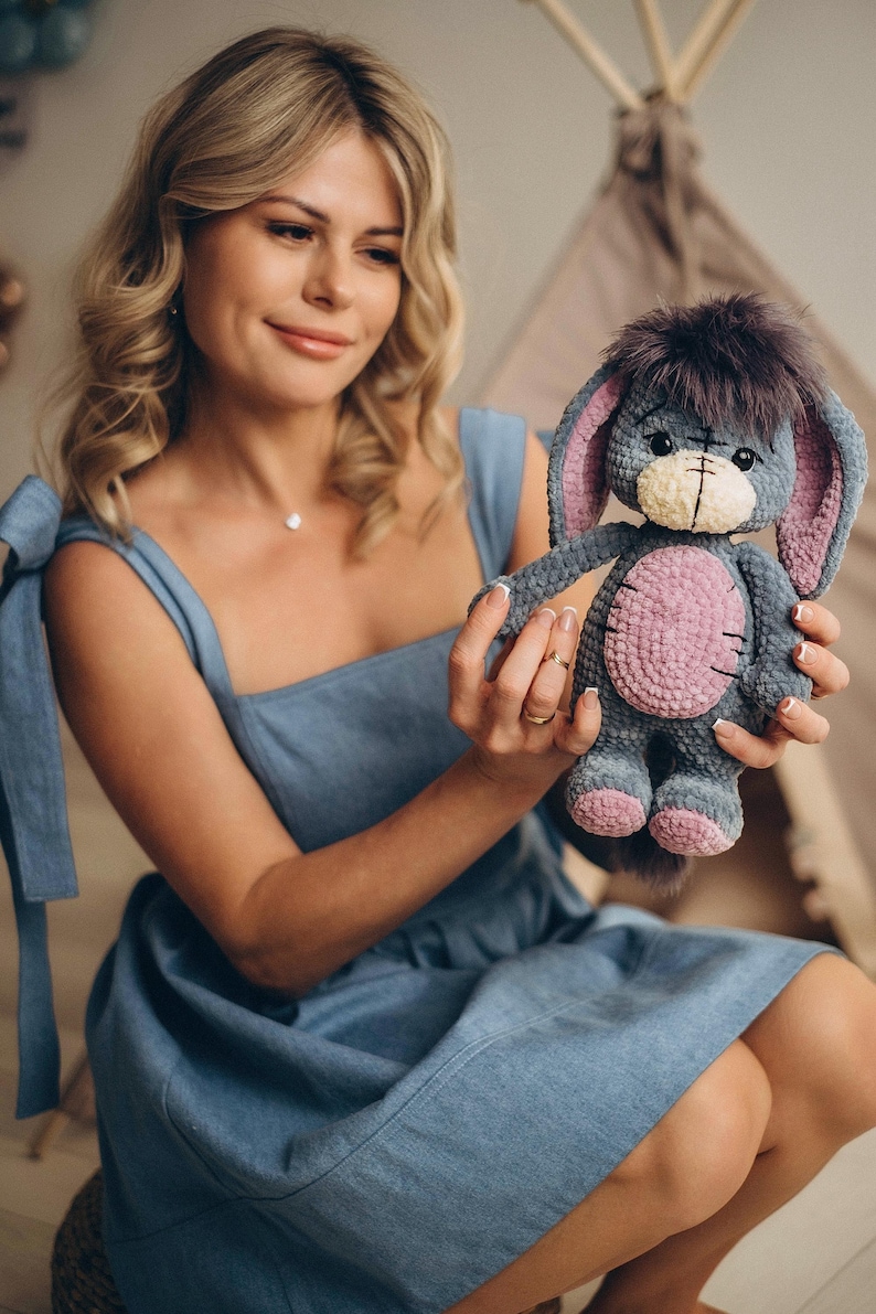 Pu&ograve; includere: Una donna con un vestito blu tiene in mano un peluche di stoffa grigio e rosa lavorato a uncinetto con orecchie lunghe. L'animale ha un viso cucito con occhi neri e un naso nero.