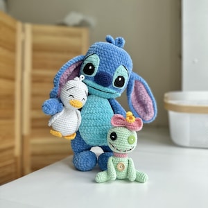 Puede incluir: Una criatura azul de ganchillo con orejas grandes y vientre azul claro, sosteniendo un pequeño pato blanco con pico naranja. A su lado hay una muñeca de ganchillo verde claro con un lazo rosa, un botón en el pecho y ojos de botón desiguales.