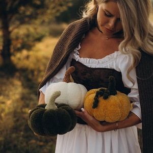 Puede incluir: Una persona sostiene tres calabazas de peluche en tonos verde, blanco y amarillo. Las calabazas tienen superficies texturizadas y tallos verdes o marrones. La persona lleva un vestido blanco y un suéter marrón.