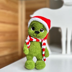 Peut inclure: Une peluche crochet&eacute;e verte &agrave; la texture douce, dot&eacute;e de grands yeux sombres, d'un nez brod&eacute; noir et d'une petite bouche. La figurine porte un bonnet de P&egrave;re No&euml;l rouge vif avec un bord blanc et une &eacute;charpe ray&eacute;e rouge et blanche assortie.