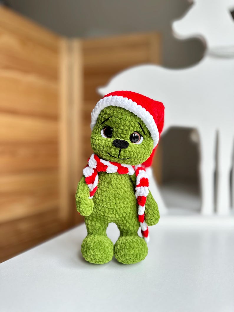 Peut inclure: Une peluche crochet&eacute;e verte &agrave; la texture douce, dot&eacute;e de grands yeux sombres, d'un nez brod&eacute; noir et d'une petite bouche. La figurine porte un bonnet de P&egrave;re No&euml;l rouge vif avec un bord blanc et une &eacute;charpe ray&eacute;e rouge et blanche assortie.