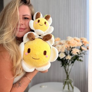 K&ouml;nnte beinhalten: Zwei l&auml;chelnde, pl&uuml;schige blumenf&ouml;rmige Spielzeuge. Das gr&ouml;&szlig;ere wird vor dem kleineren gehalten. Die Blumen sind gelb mit wei&szlig;en Bl&uuml;tenbl&auml;ttern und braunen Akzenten. Der Hintergrund umfasst eine Vase mit Blumen und einen wei&szlig;en Tisch.