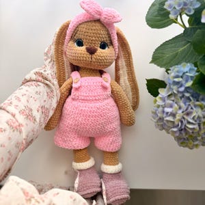 May include: A crocheted rabbit doll with tan yarn fur, wearing a pink headband with a bow, pink overalls with small buttons, and mauve-colored boots with white cuffs. The rabbit has long ears, embroidered facial features, and blue eyes.