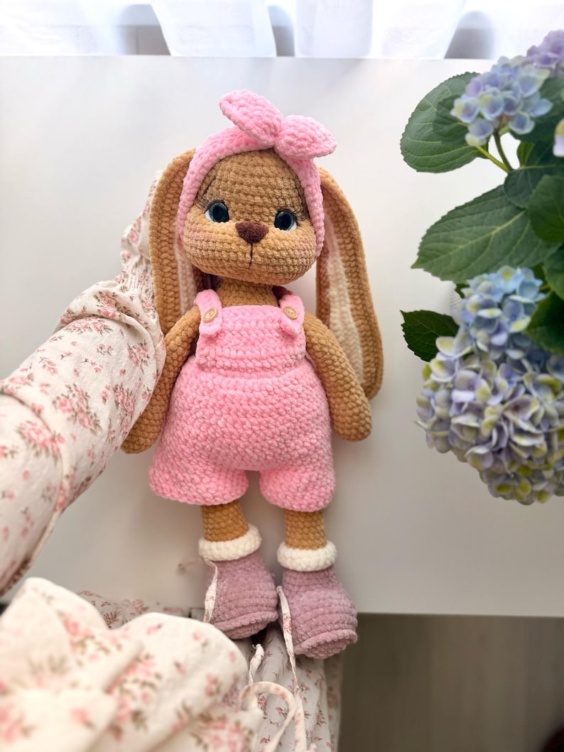 May include: A crocheted rabbit doll with tan yarn fur, wearing a pink headband with a bow, pink overalls with small buttons, and mauve-colored boots with white cuffs. The rabbit has long ears, embroidered facial features, and blue eyes.