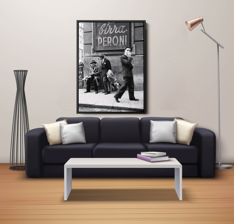 May include: A black and white framed print of a vintage street scene with the text "birra PERONI". The print hangs above a black sofa with white pillows and a white coffee table. A modern floor lamp and decorative black metal sculpture are also in the room.