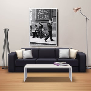 May include: Black and white framed print of a vintage street scene. The image features men in suits, a sign that reads "Birra Peroni", and a dog. The print is displayed above a black sofa with white pillows.