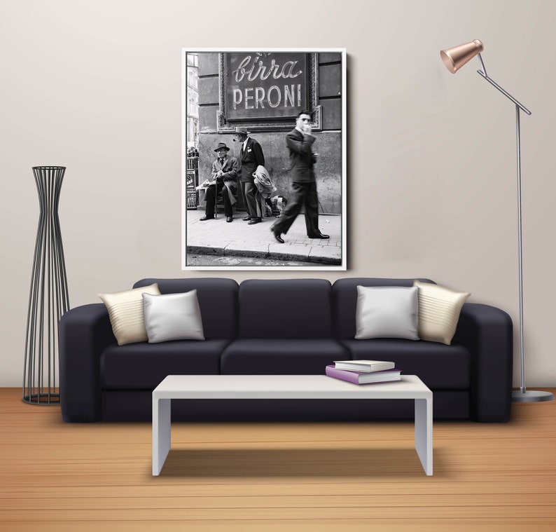 May include: Black and white framed print of a vintage street scene. The image features men in suits, a sign that reads "Birra Peroni", and a dog. The print is displayed above a black sofa with white pillows.