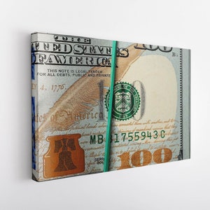 May include: A bundle of US $100 bills held together with a green rubber band. The bills are face down and the text "THE UNITED STATES OF AMERICA" is visible on the top bill. The text "THIS NOTE IS LEGAL TENDER FOR ALL DEBTS, PUBLIC AND PRIVATE" is also visible. The serial number "MB 317559430" is visible on the bottom bill.