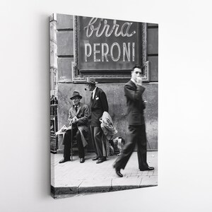 May include: Black and white photograph of a street scene featuring three men near a sign that reads "Birra Peroni". One man smokes a pipe, another sits on a bench, and a third walks by smoking a cigarette. The image is a vintage style print.