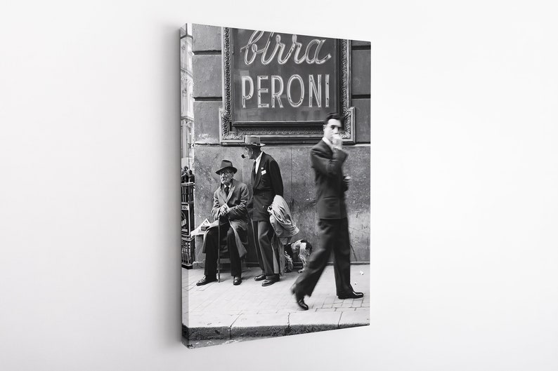 May include: Black and white photograph of a street scene featuring three men near a sign that reads "Birra Peroni". One man smokes a pipe, another sits on a bench, and a third walks by smoking a cigarette. The image is a vintage style print.