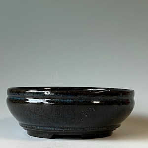 May include: A round, shallow ceramic bowl with a dark, glossy glaze featuring deep blue and black tones. The bowl has a smooth, curved exterior with a subtle horizontal ridge near the rim and a raised circular base, showcasing a reflective finish.