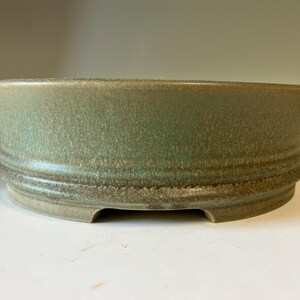 May include: A round, shallow ceramic bonsai pot with a textured, mottled green and brown glaze. The exterior features a raised horizontal band near the base, and the bottom edge has small, integrated feet that create a slight gap from the surface.