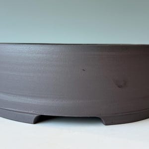 May include: A gray ceramic bonsai pot with a rounded shape. The pot has a matte finish and a low profile, with a slightly curved rim and a small footed base. The pot is designed for planting small trees.