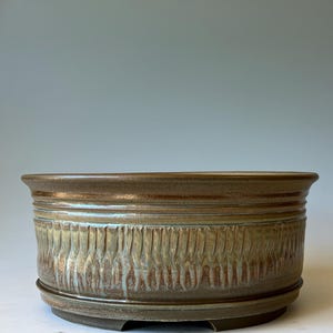 May include: A round, low-profile ceramic bonsai pot with a wide, flared rim. The exterior features a textured, vertical ribbed pattern finished in a variegated glaze of earthy brown, muted green, and subtle blue tones with a smooth base.