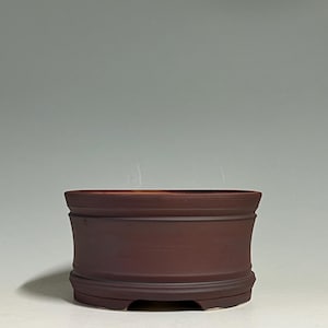 May include: A dark brown, cylindrical ceramic bonsai pot with a smooth, matte finish. The pot features a slightly flared rim and a raised base with small, arched cutouts along the bottom edge, providing a stable and functional design for planting.