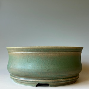 May include: A round, low-profile ceramic bonsai pot with a matte, sage green glaze. The vessel features a slightly flared rim, a curved body, and a raised base with small cutouts along the bottom edge, showcasing a textured, earthy finish.
