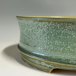 May include: A shallow, circular ceramic bonsai pot featuring a textured, mottled green glaze with white crystalline speckling. The exterior wall curves slightly outward toward the rim, and the base includes small, integrated feet for elevation.