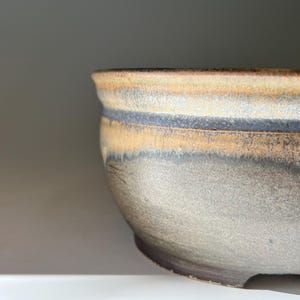 May include: A ceramic bowl featuring a rounded silhouette with a thick, flared rim. The exterior displays a layered glaze effect, transitioning from a warm, sandy beige at the top to a muted, cool gray and dark charcoal tone toward the base.