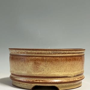 May include: A round ceramic bonsai pot with a glazed finish in shades of tan, beige, and brown. The exterior features a horizontal band design with a slightly raised rim and base. The pot has a wide, open top and a footed bottom with cutouts.