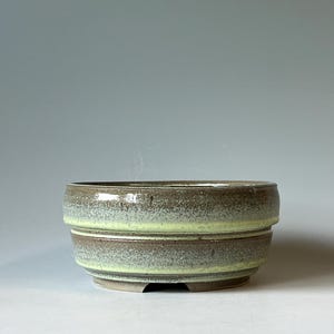 May include: A round ceramic bowl with a stepped exterior design. The glaze features a mottled brown and gray texture with a prominent horizontal band of bright lime green encircling the middle section of the vessel's exterior wall.