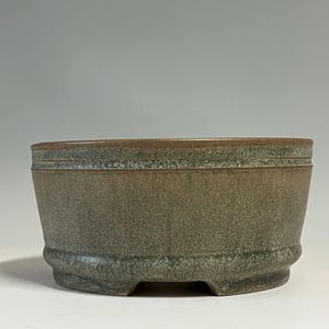 May include: A round ceramic bonsai pot with a textured, mottled finish in shades of muted green and brown. The vessel features a slightly flared rim, a horizontal incised line detail near the top, and a sturdy base with small cut-out feet.